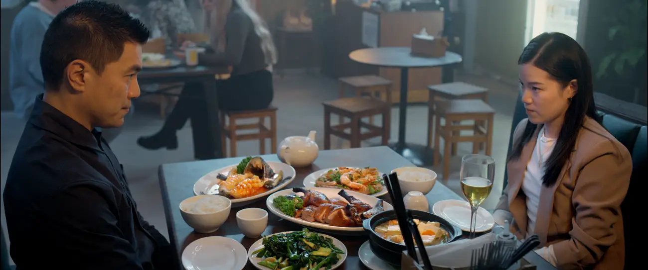 Winson and Elle's look at each other unhappily across a table full of food in a Chinese diner.
