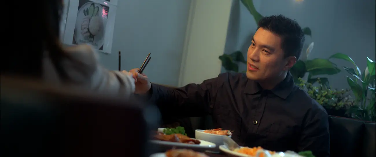 Dutch angle shot of Winson confidently deflecting chopsticks over a table full of food.