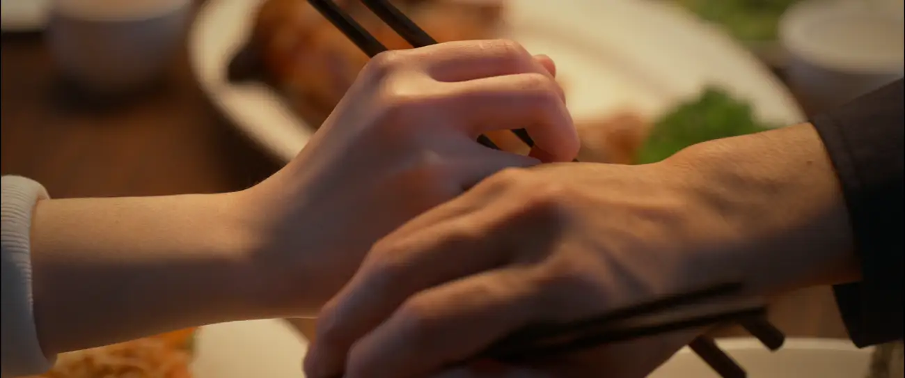 A close up of man and woman's hands touching while reaching out for food with their chopsticks.