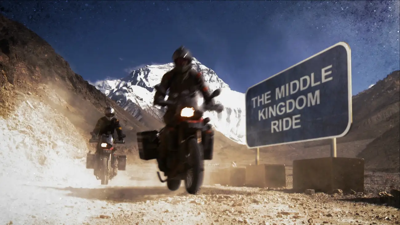 Two motorcyclists drive past a roadsign saying 'The Middle Kingdom Ride' on a mountainous dirt road.
