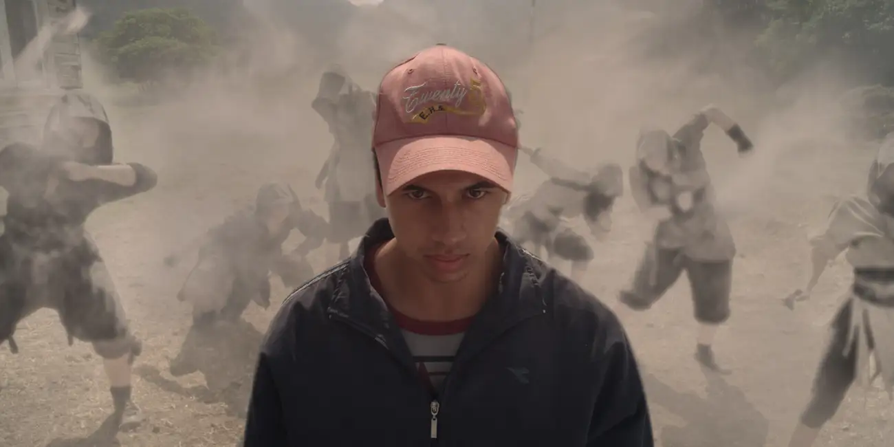 A young Māori man with shadowy figures appearing in a dust cloud behind him.
