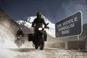Two motorcyclists drive past a roadsign saying 'The Middle Kingdom Ride' on a mountainous dirt road.