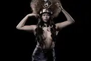 A half-naked Samoan woman steadies a large traditional headdress on her head, with the 'Teine Sā: The Ancient Ones' title graphic.